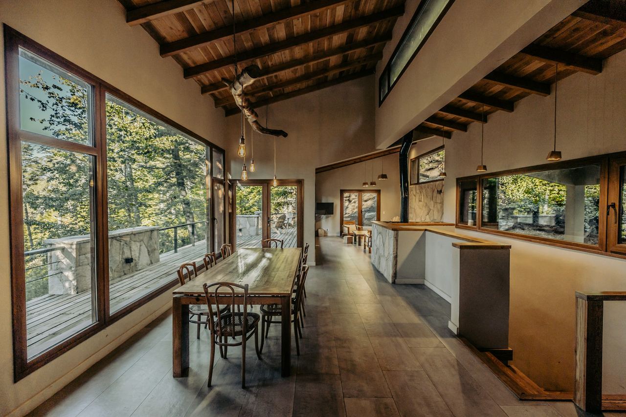 Warm mountain-modern kitchen and dining room with tall windows facing a pine forest